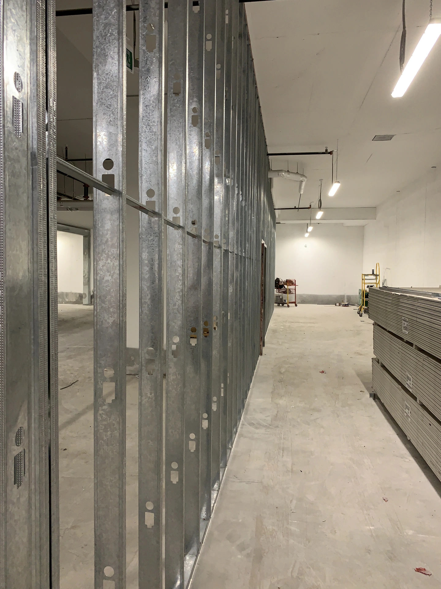 A large, unfinished room with metal wall studs installed, stacks of drywall sheets on the right, and exposed ceiling fixtures.