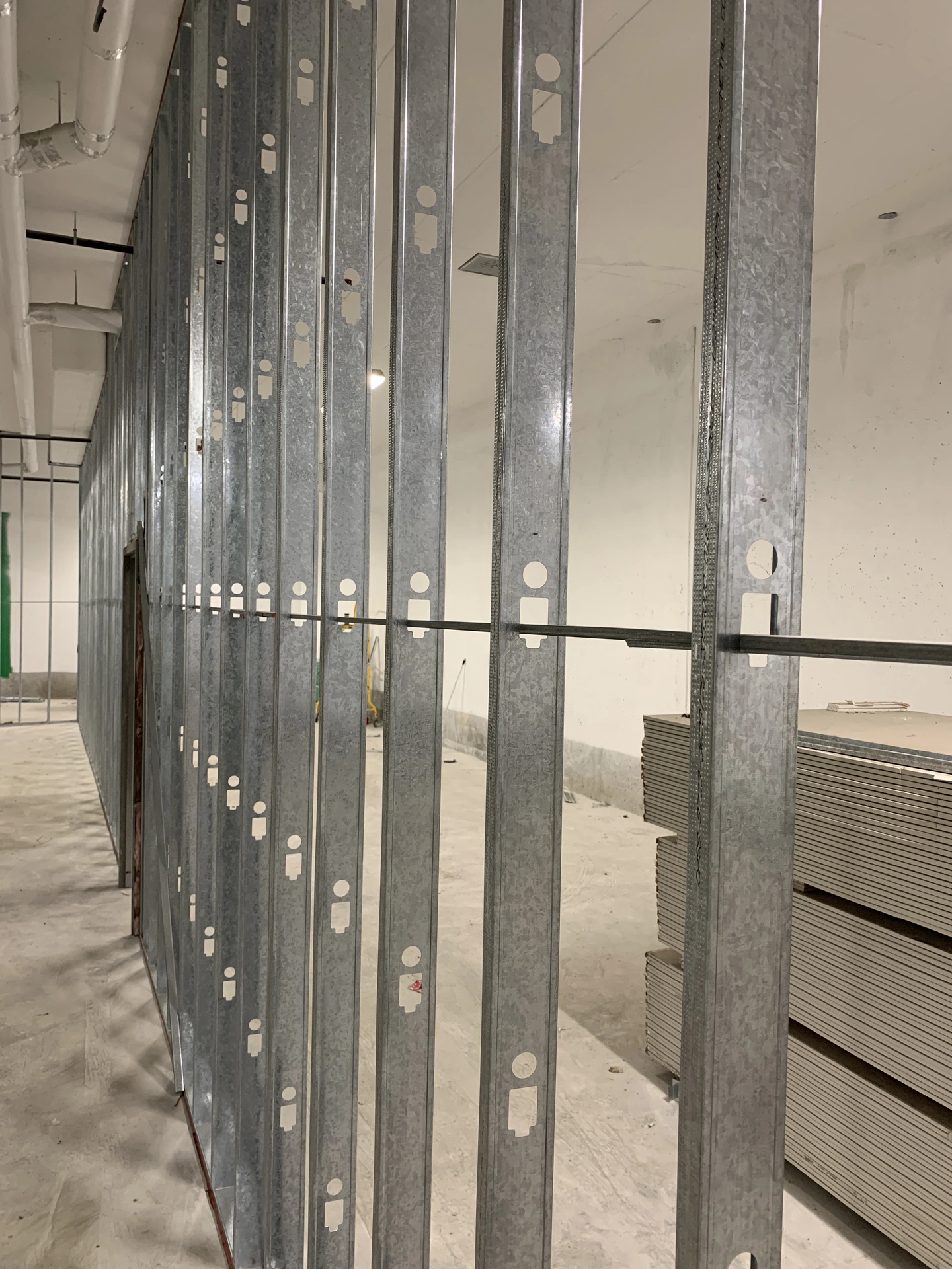 A large, unfinished room with metal wall studs installed, stacks of drywall sheets on the other side of the wall