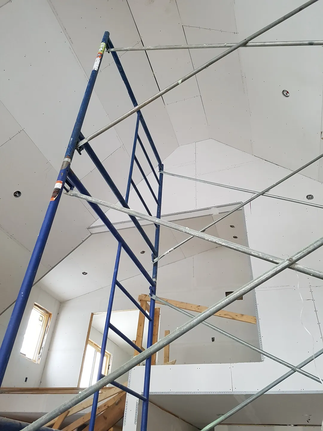 Interior of a partially constructed room with high slanted ceiling, wooden paneling on one wall, large windows, and construction materials on the floor.