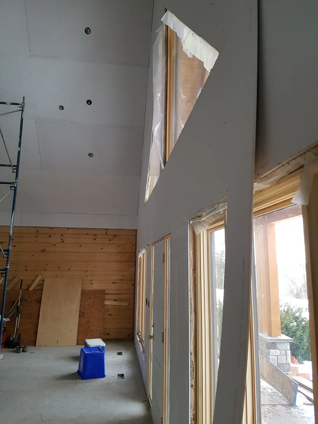 Interior of house under construction with drywall installed on walls and ceiling, scaffolding, wooden stairs, and construction materials on the floor.