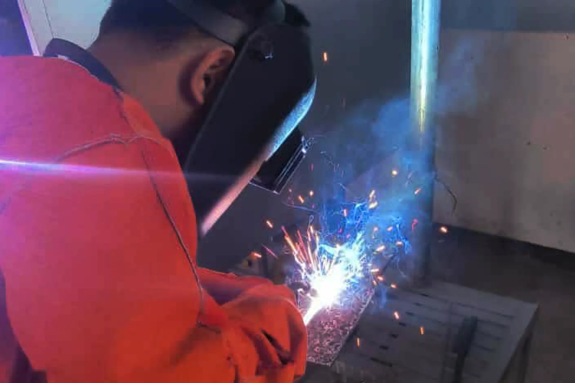 Welder wearing a protective mask and gear performing arc welding on metal with sparks flying
