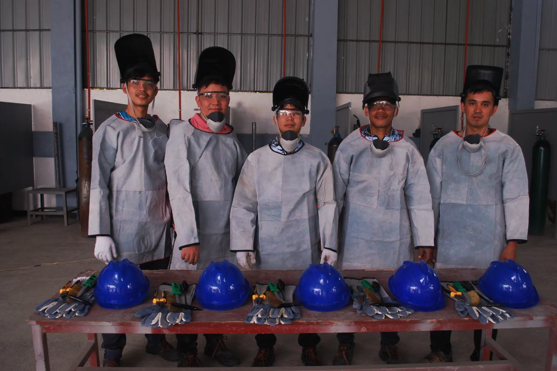 A group of trainee welders in protective aprons and masks standing behind a table with blue hard hats and gloves