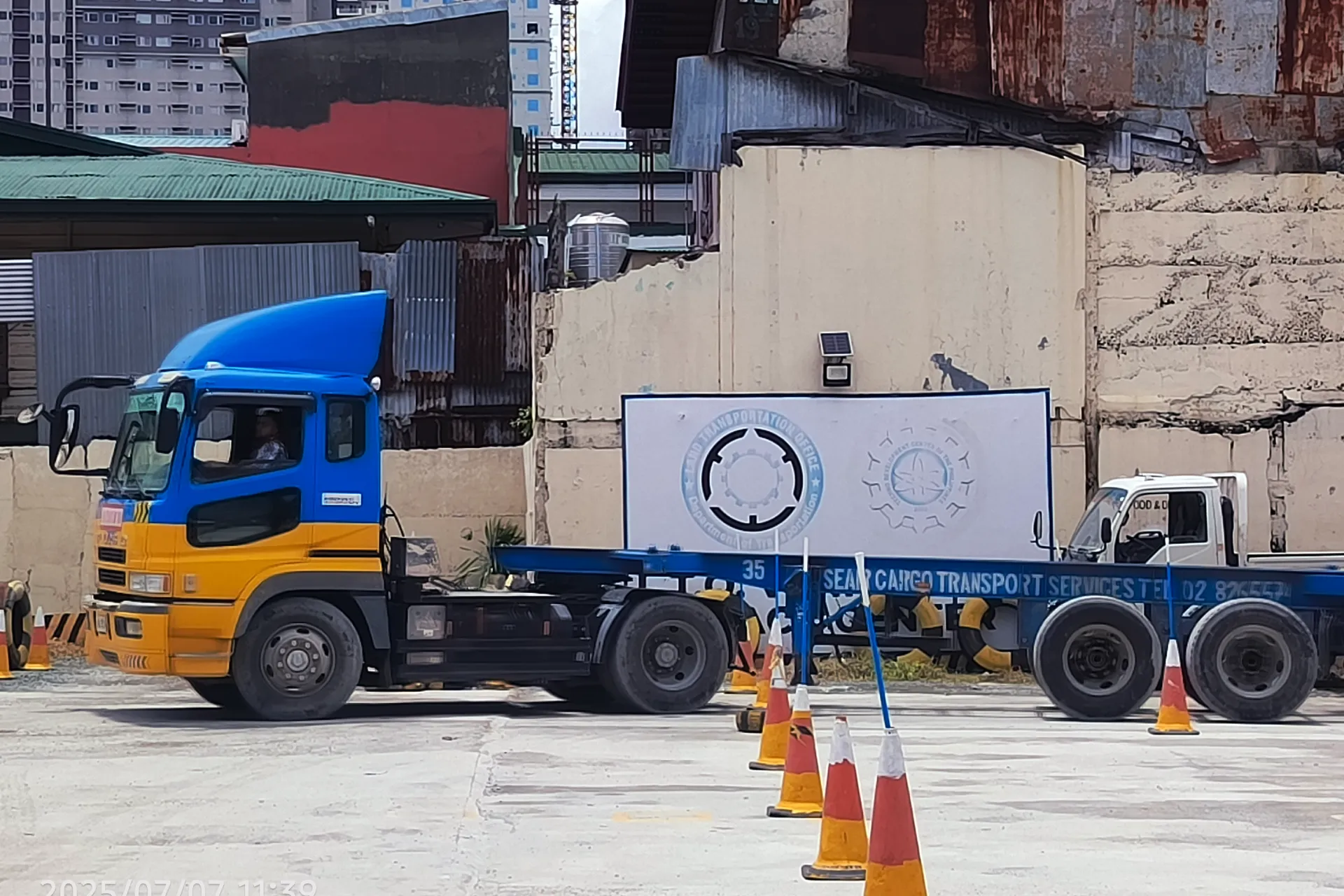 Blue and yellow heavy training truck parked at the LTO vehicle testing center with a large logo in the background