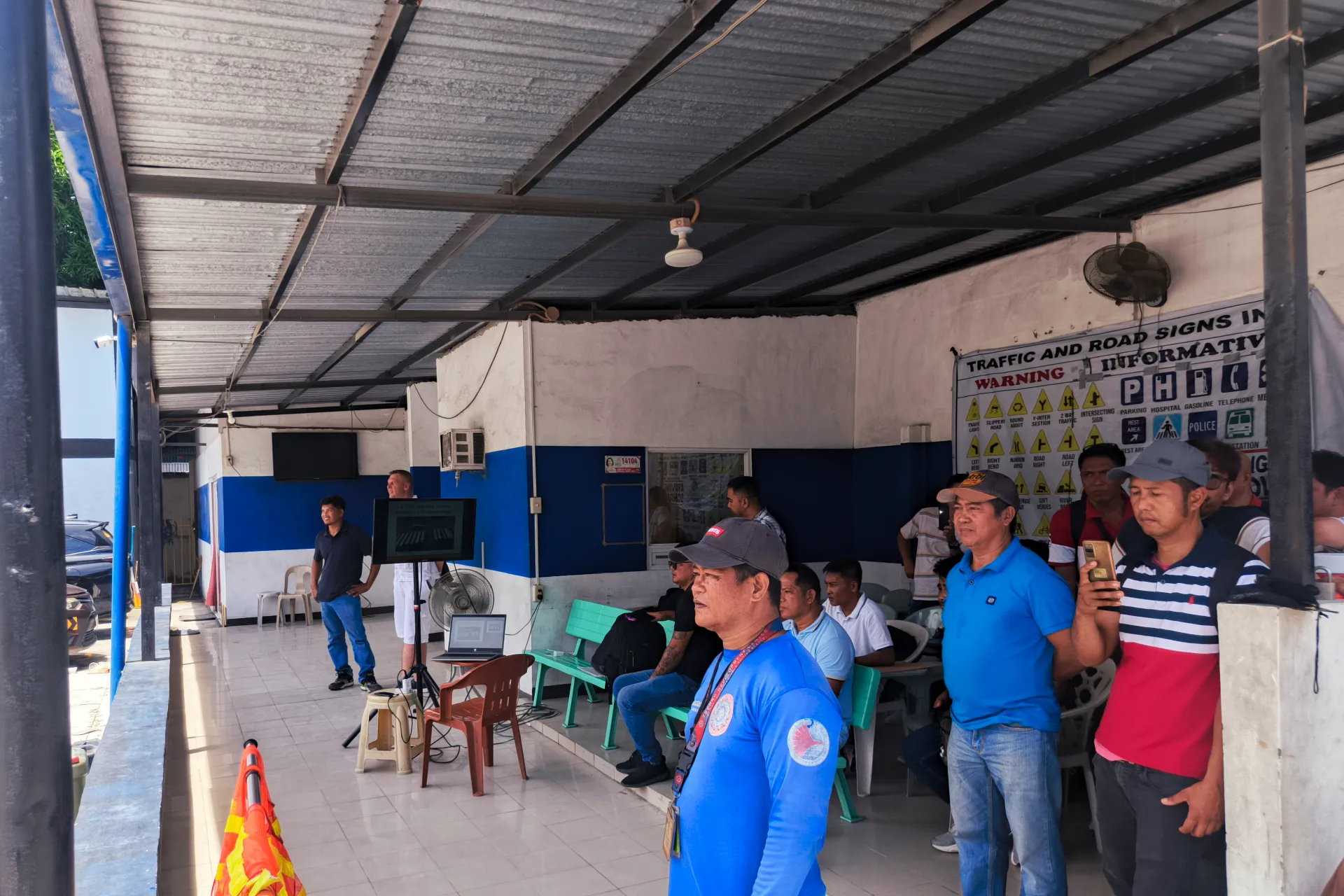 Students attending a driving theory briefing in an open-air classroom next to a large poster of Philippine traffic and road signs.