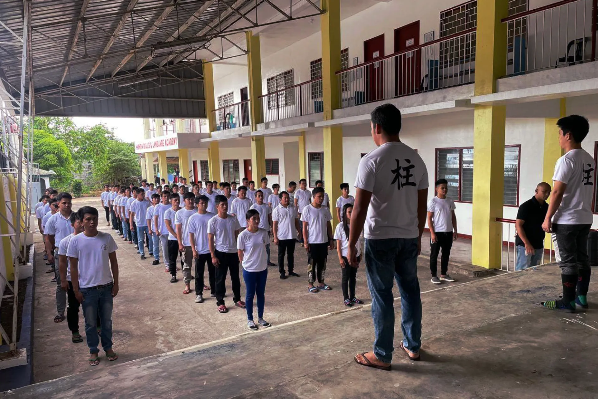 A large group of trainees in white t-shirts standing in formation during an outdoor assembly being addressed by an instructor at Hashira Academy