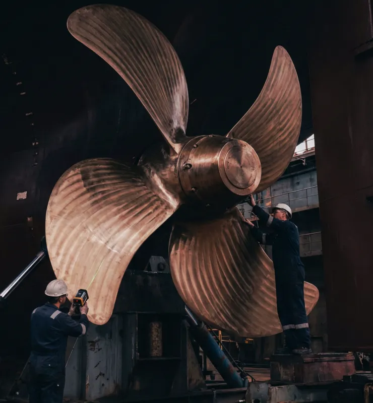 Shipyard workers inspecting a massive bronze ship propeller in a dry dock facility.