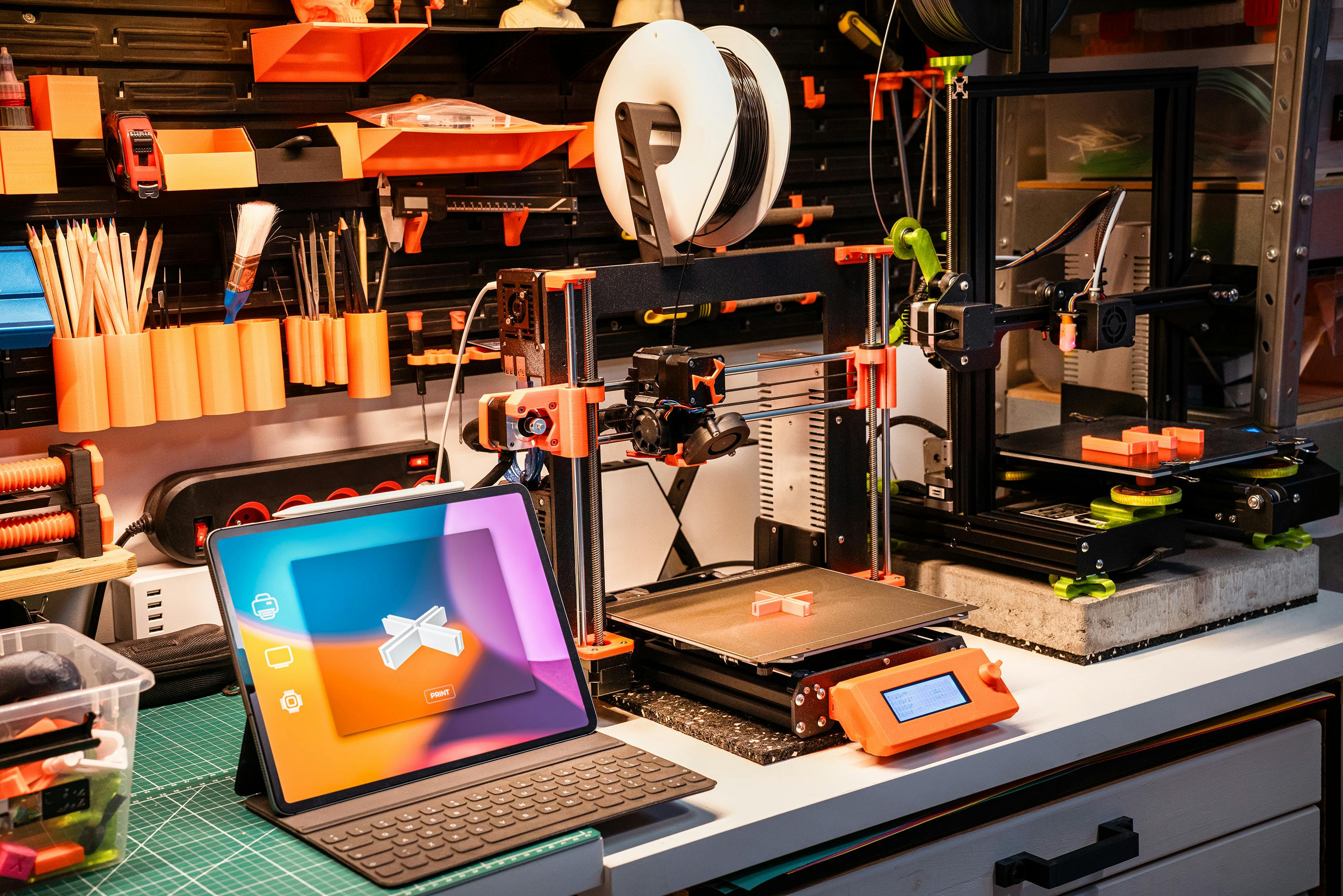 Workspace with two 3D printers actively printing orange objects and a tablet displaying a 3D print design on a cutting mat.