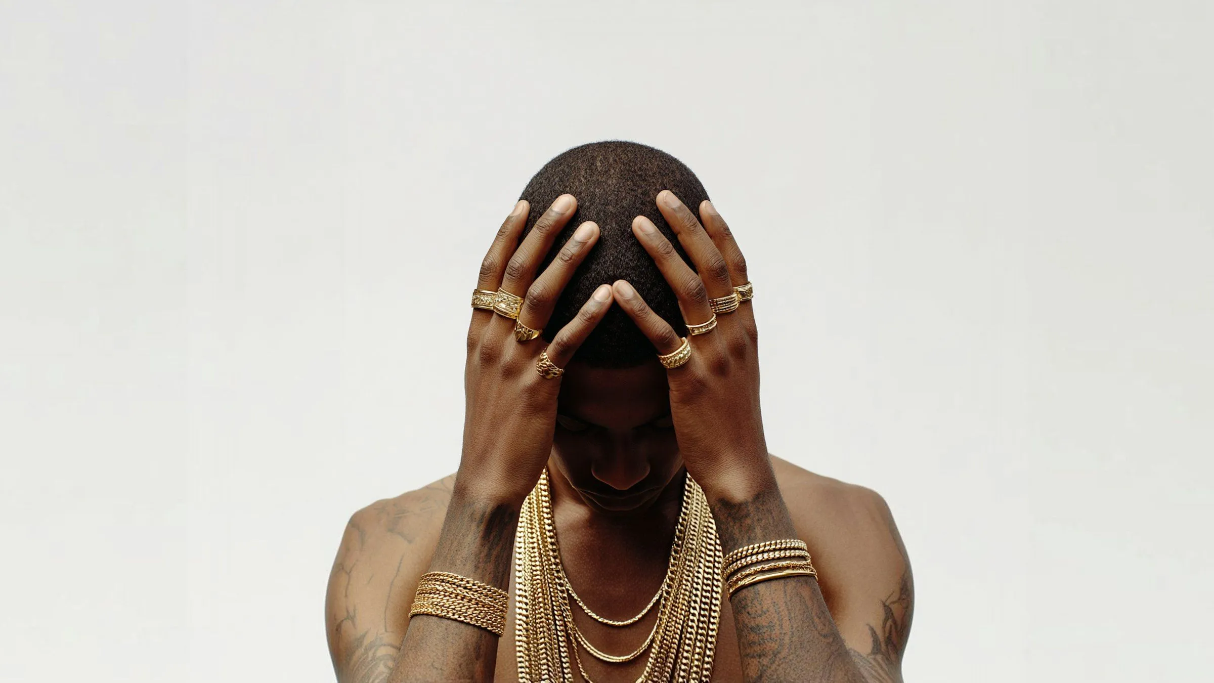 Person with dark skin and short hair wearing multiple gold rings, bracelets, and layered gold necklaces, holding their head with both hands.