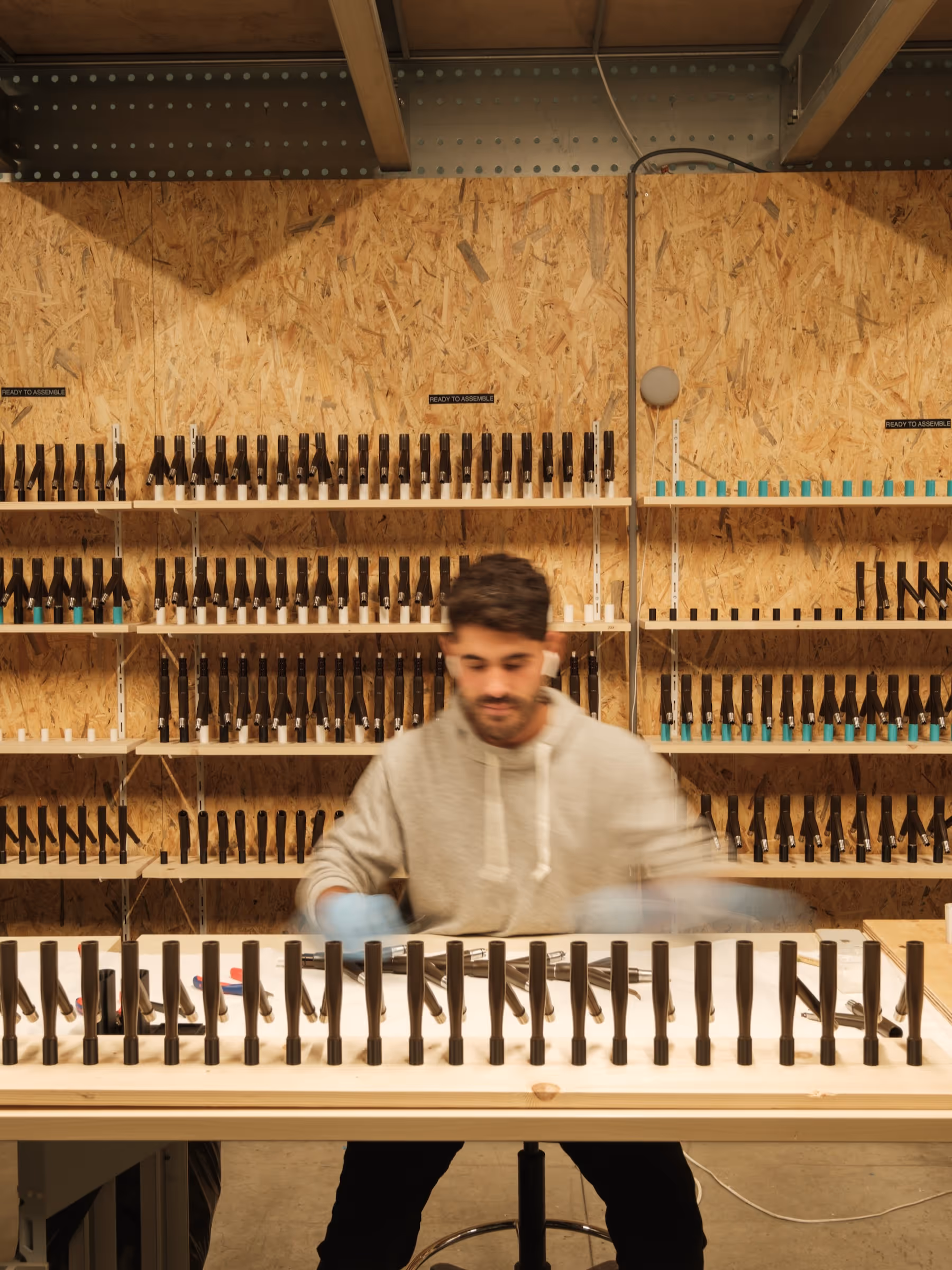 Person wearing gloves assembling black metal parts on a table in a workshop with shelves full of similar parts.