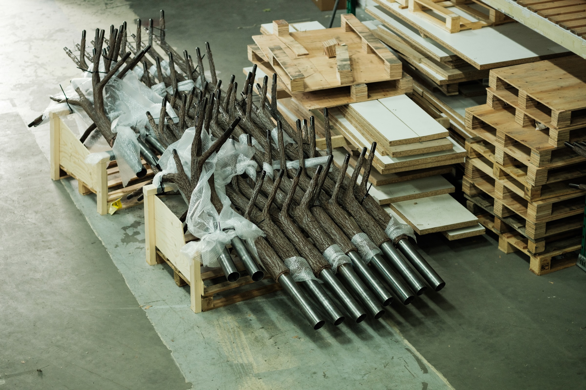Stacked artificial tree trunks with metal bases wrapped in bubble wrap in a warehouse next to wooden pallets and boards.