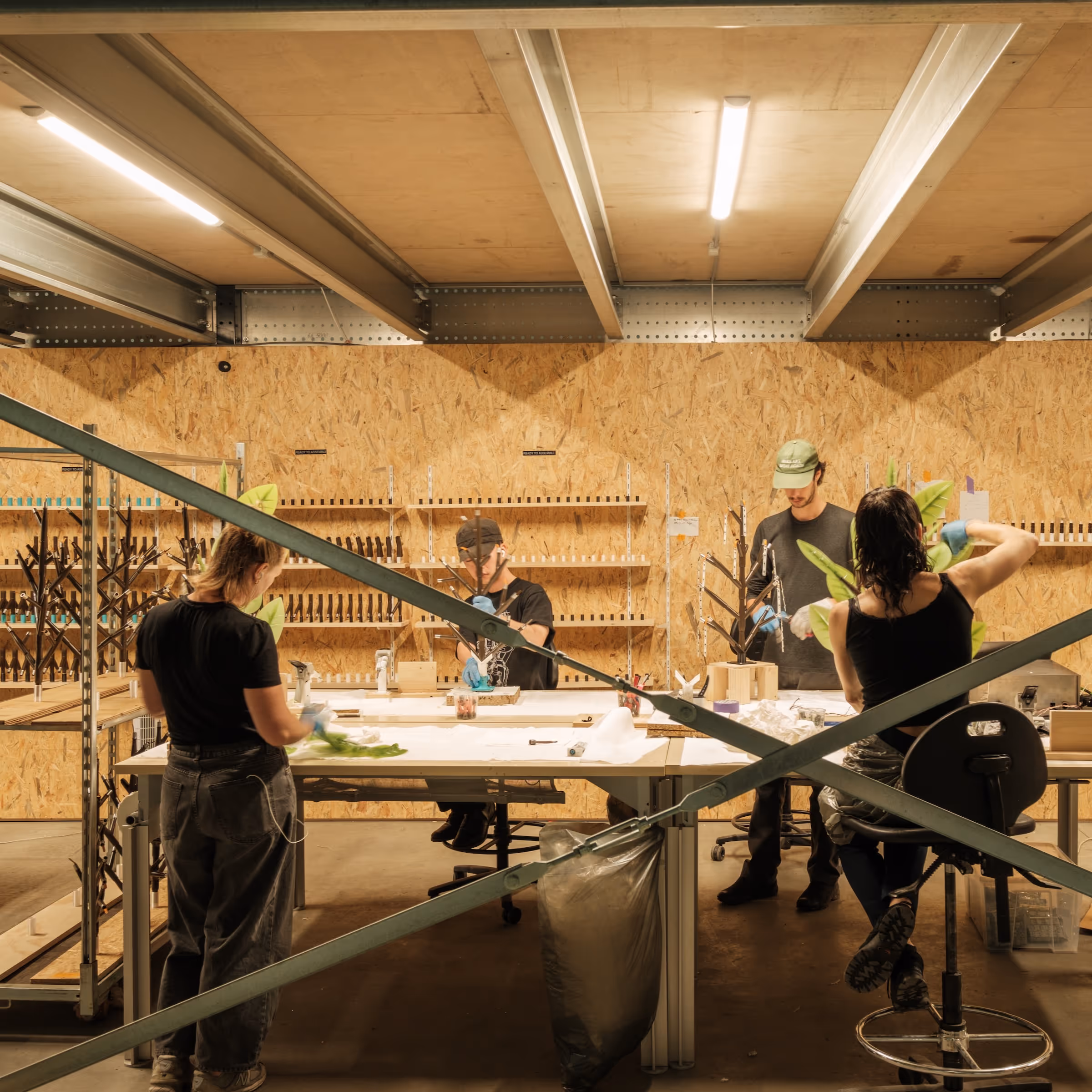 Four people assembling products at workbenches in a well-lit industrial workshop with wooden walls.