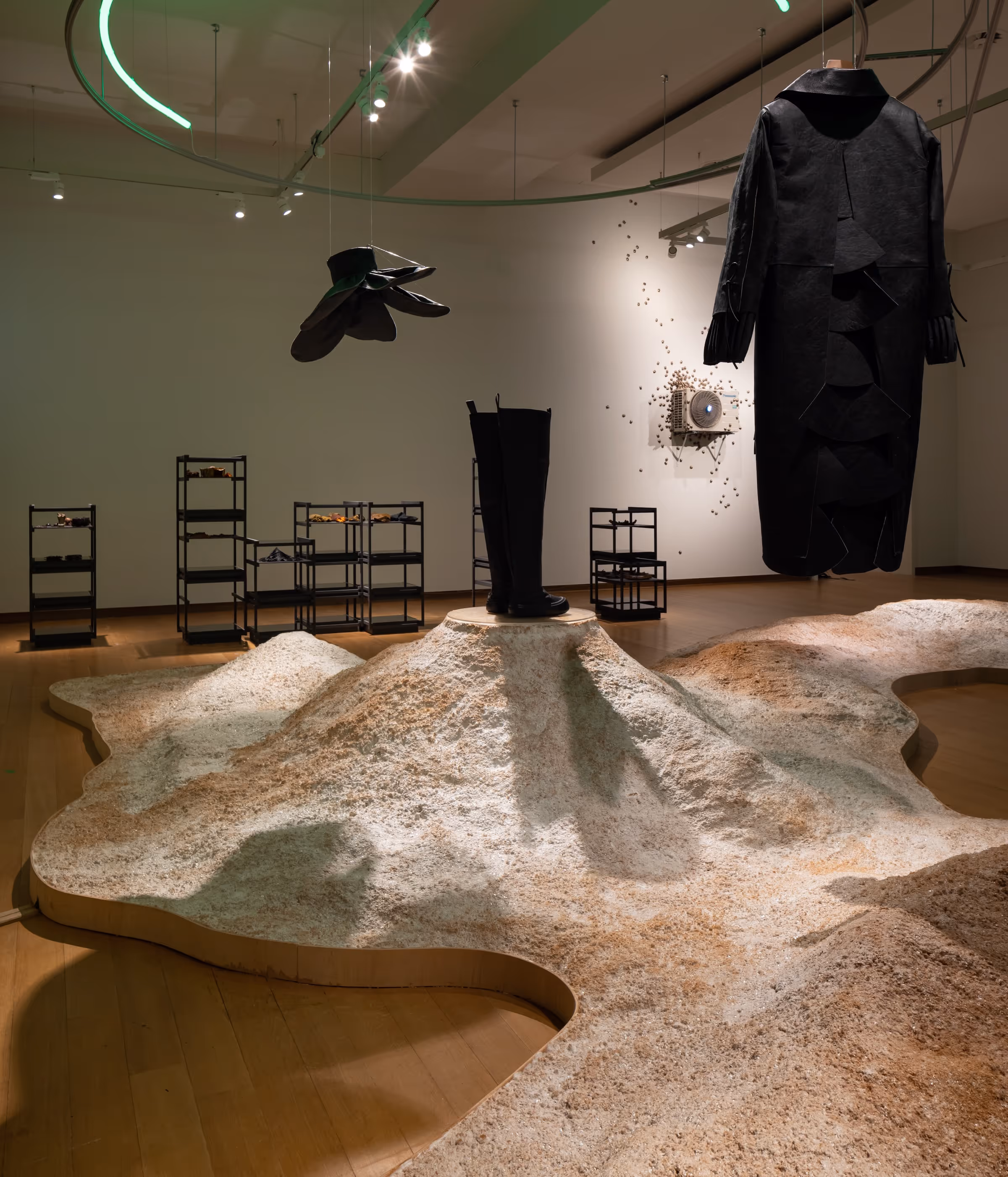 Contemporary art installation with a large black boot on a mound, a hanging black coat, suspended black gloves, mycelium floor installation on a gallery floor.