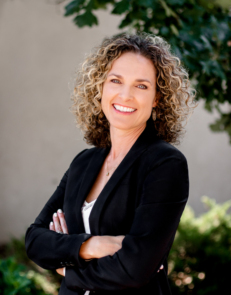 Amilym Stange with curly hair wearing a black blazer standing outdoors with arms crossed, llooking professional.