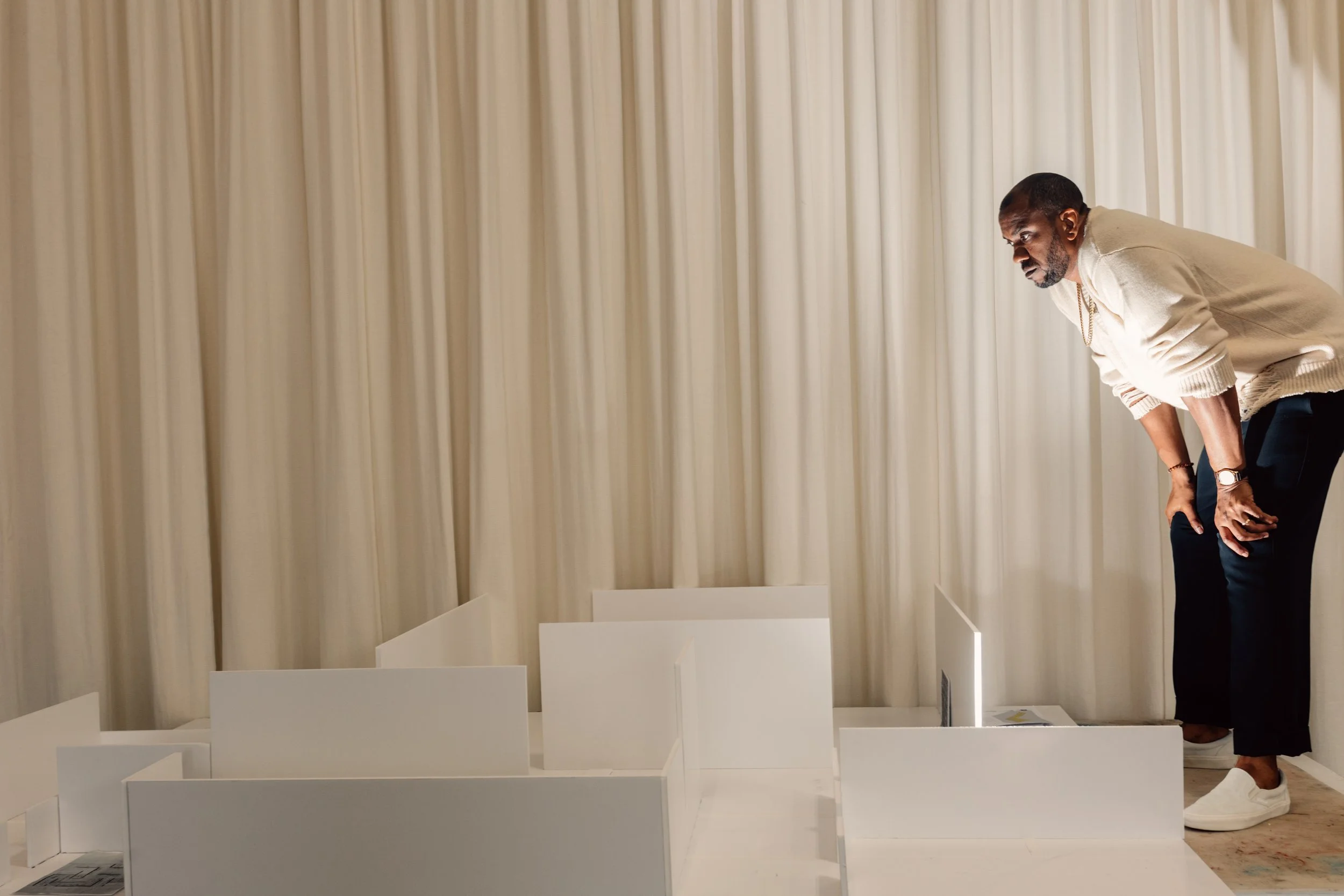A bearded Black man stands in front of undulating white curtains. He bends slightly, hands on knees, intensely surveying a large in-progress diorama made of white cardboard.
