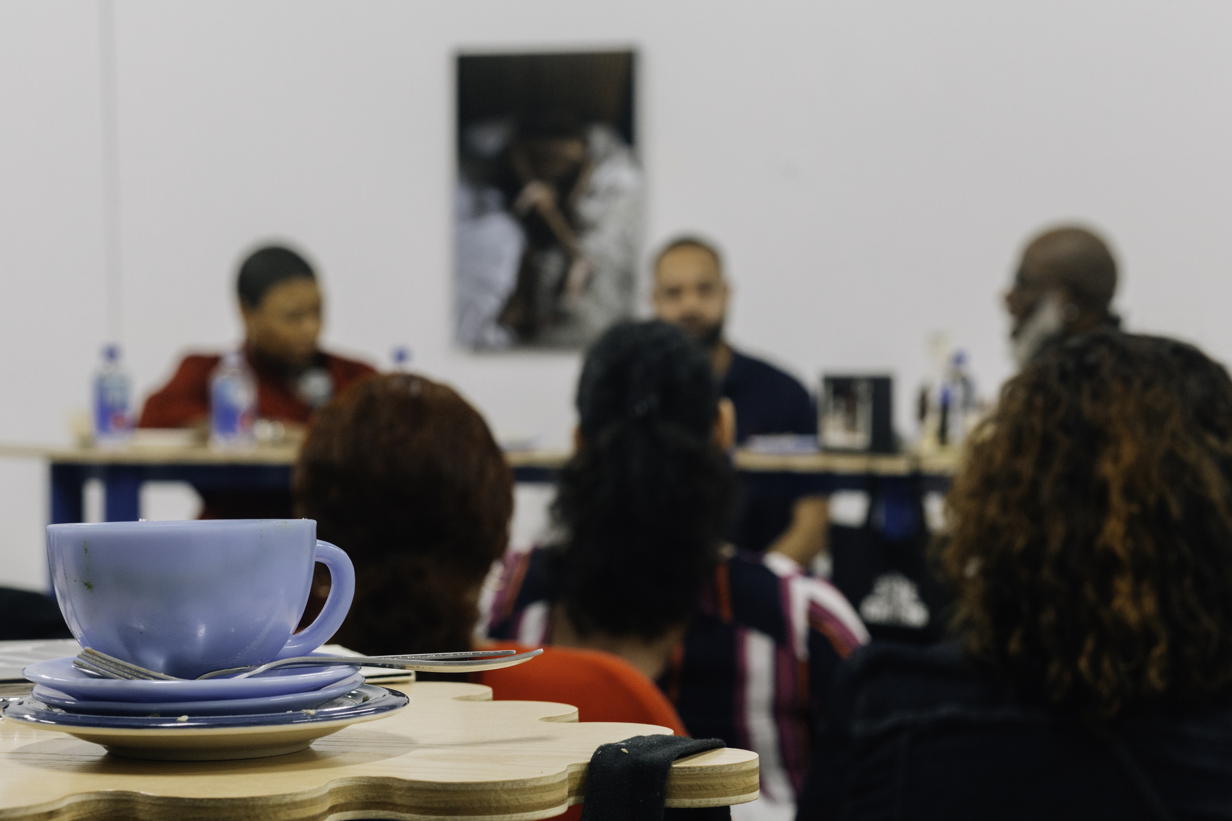A cup on saucers sits in the foreground, while in the background attendees sit listening to a panel of speakers.