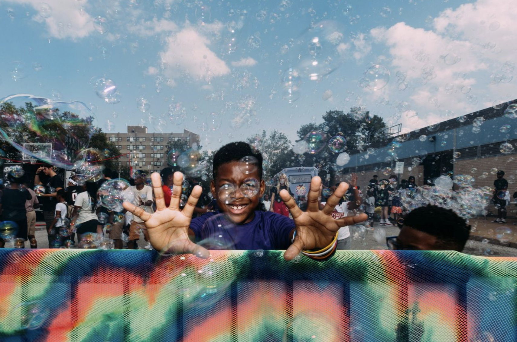 A young Black boy reaching out to the camera surrounded by bubbles, bubbles playfully ccover his eyes!