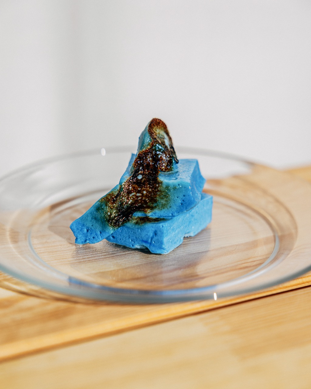 Several square cut bright blue marshmallows sit atop a modern clear glass plate. The marshmallows have been bruleed, creating a dark brown crust of burnt sugar.