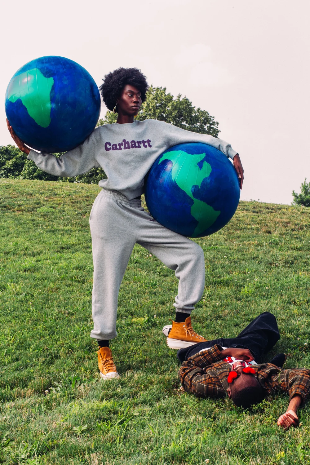 A young Black woman in grey Carhartt branded sweats stands in a power post.One foot rests on a man lying prone on the grass. She carries one giant globe under her right arm, and hoists another giant globe over her left arm. She stares defiantly off screen to the right.