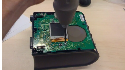 Hand using a screwdriver to assemble a green circuit board inside a black plastic casing on a wooden desk.