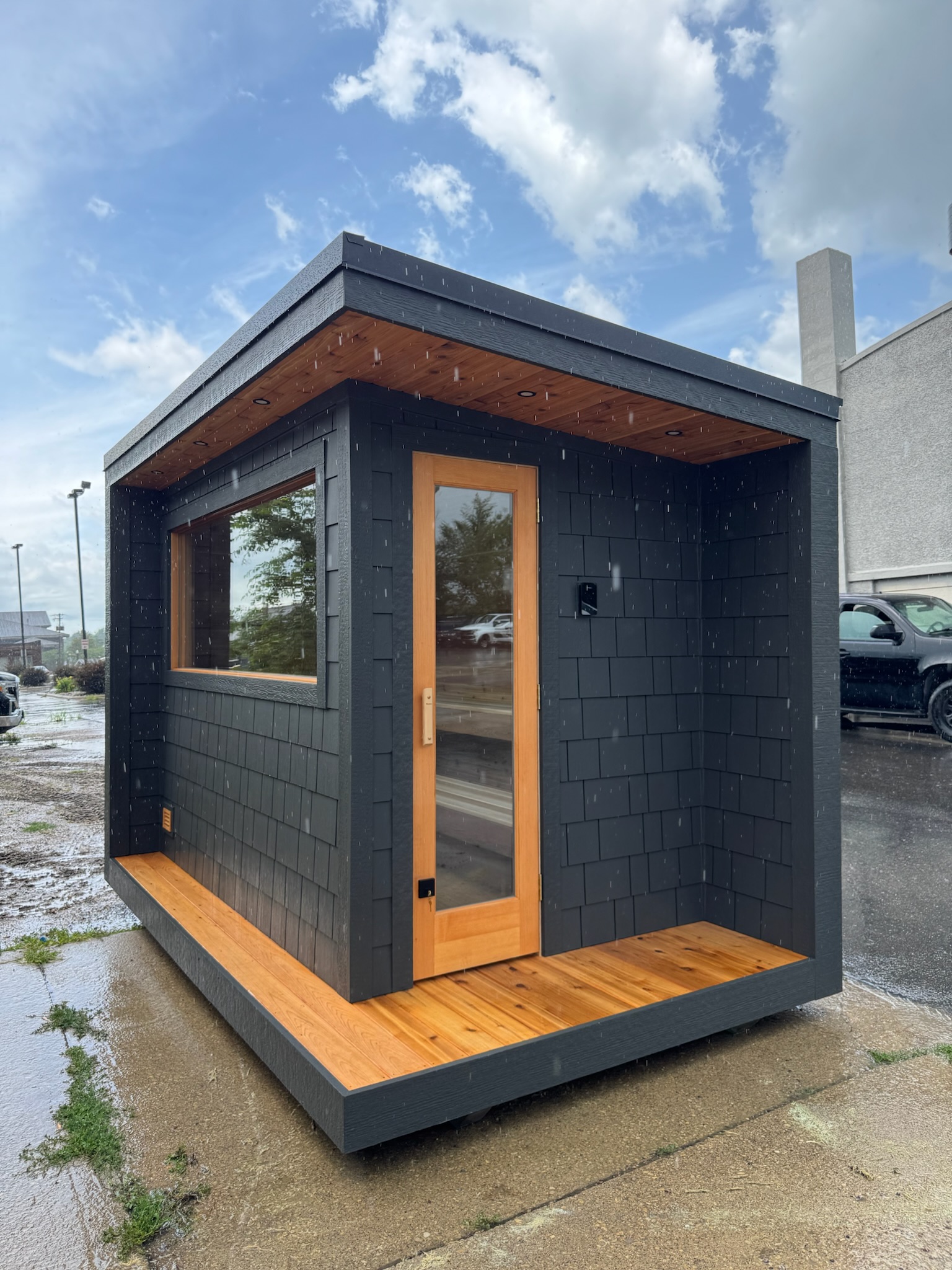 Modern small wooden sauna cabin on stone platform by a misty lake with a person swimming nearby in autumn.