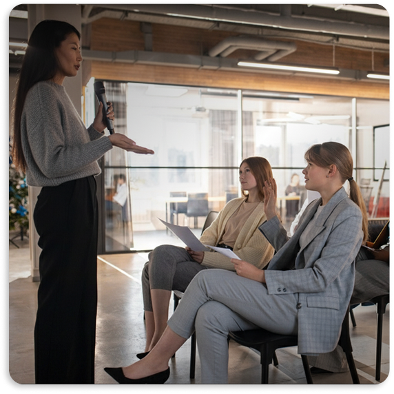 Woman standing and speaking into a microphone while two seated women listen and hold documents in a modern office setting.