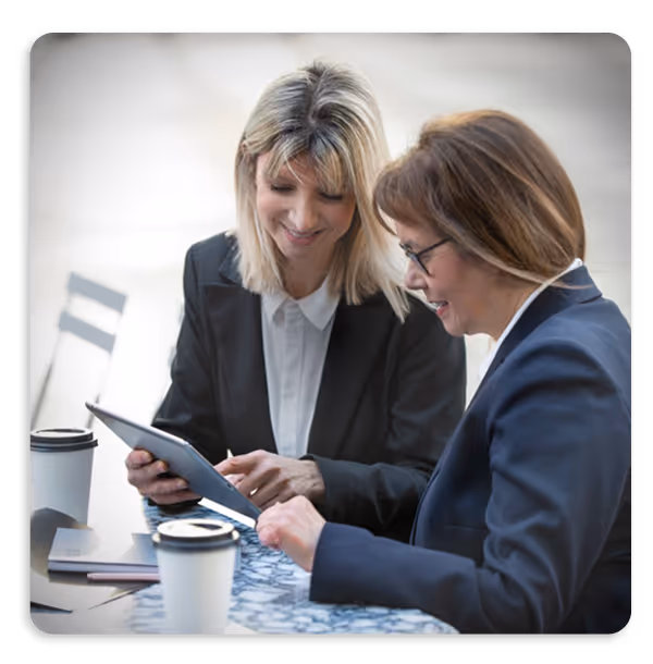 Two professional women in business attire smiling and looking at a tablet while sitting at a table with two coffee cups.