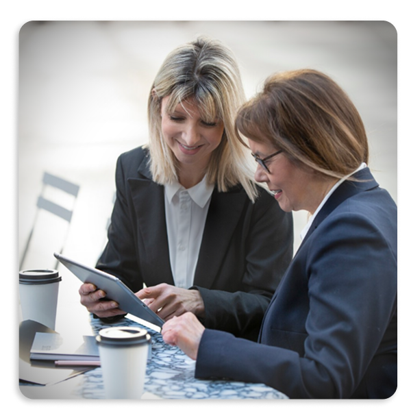 Two professional women in business attire smiling and looking at a tablet while sitting at a table with two coffee cups.