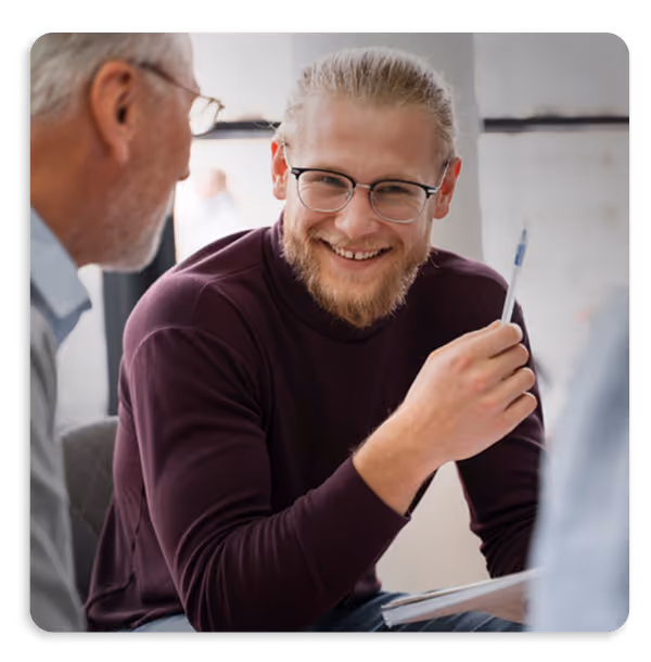 Smiling man with glasses holding a pen engaged in conversation with two other people.