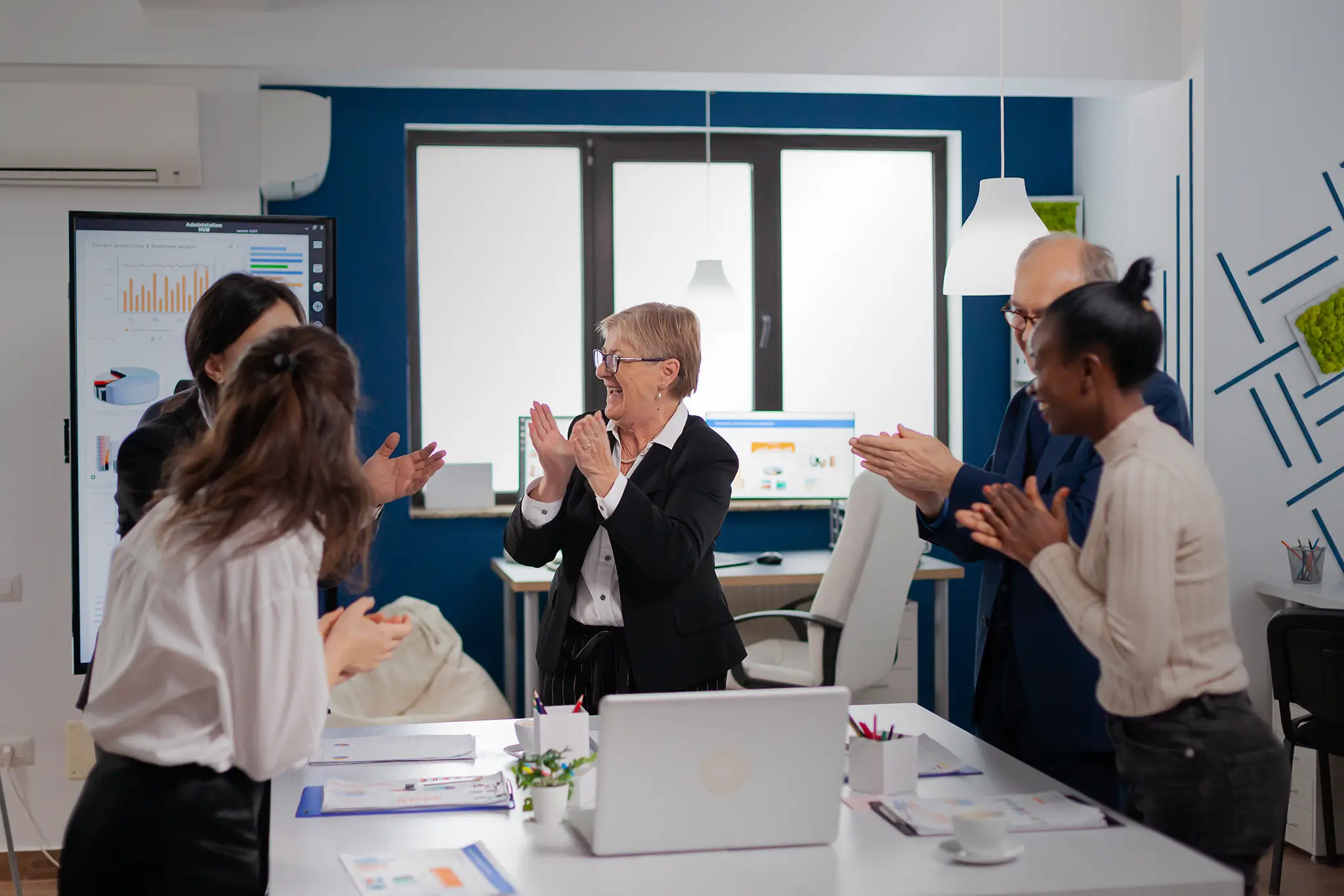 A diverse group of five professionals clapping and smiling together in a modern office meeting room.