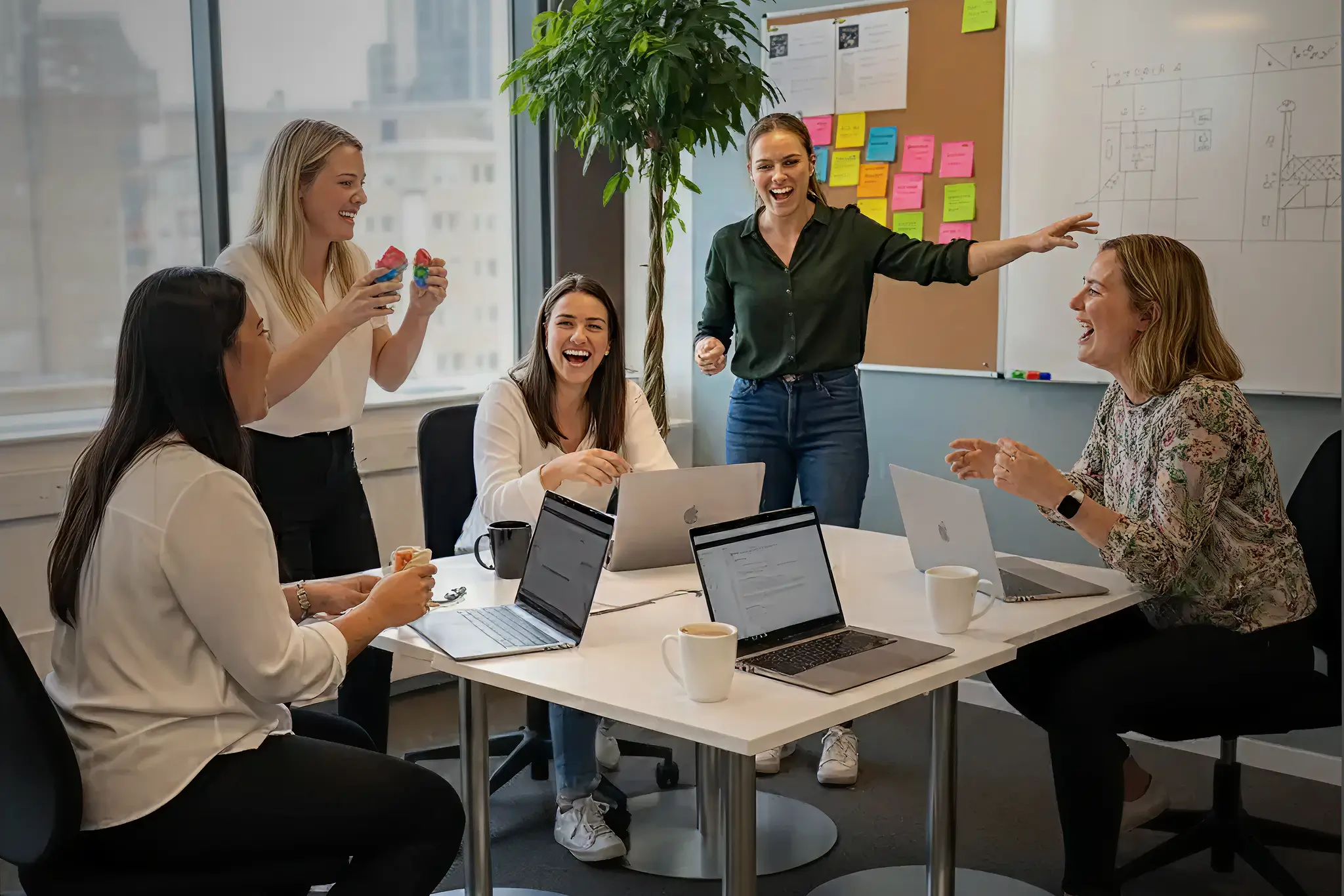 Five women laughing and interacting around a table with laptops and coffee cups in an office meeting room.