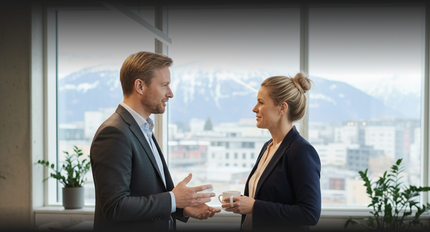 Man and woman in business attire talking and holding a coffee cup in an office with large windows overlooking a city with snowy mountains.