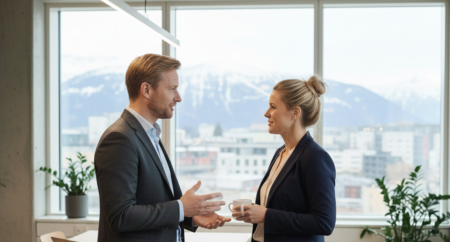 Two business professionals having a conversation in an office with large windows showing snowy mountains outside.