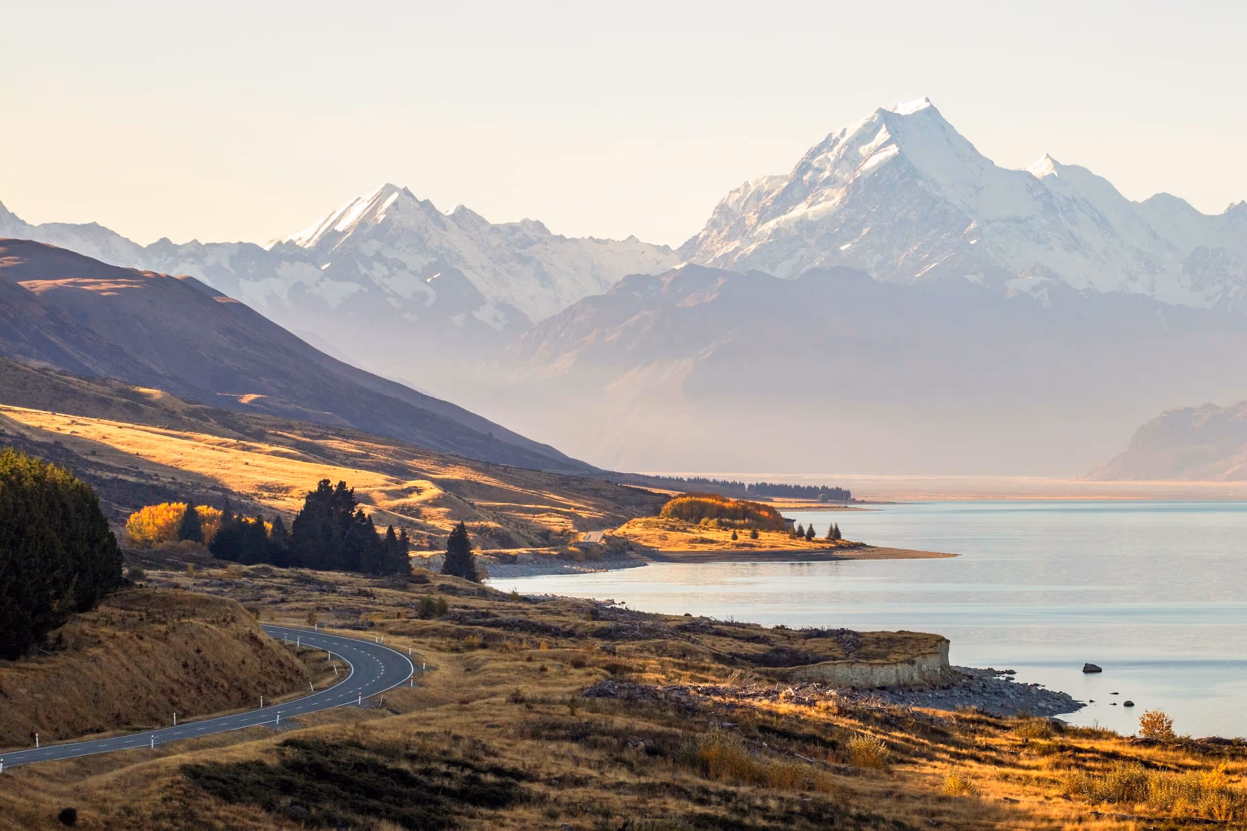 Curving road along a lakeshore with golden grass and trees, set against snow-capped mountains in the background.