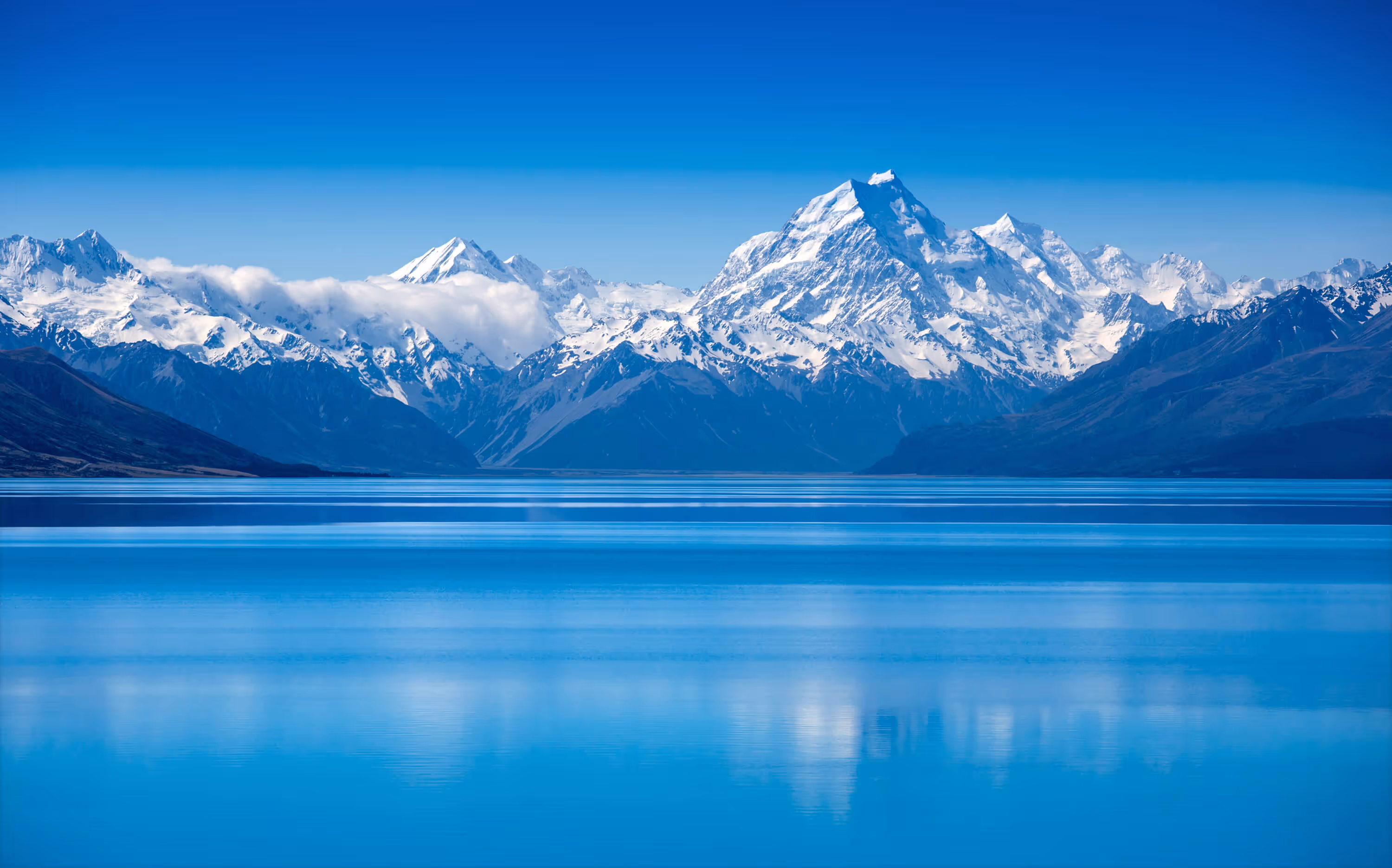Snow-covered mountain range with a clear blue sky reflected in the still water of a large lake.