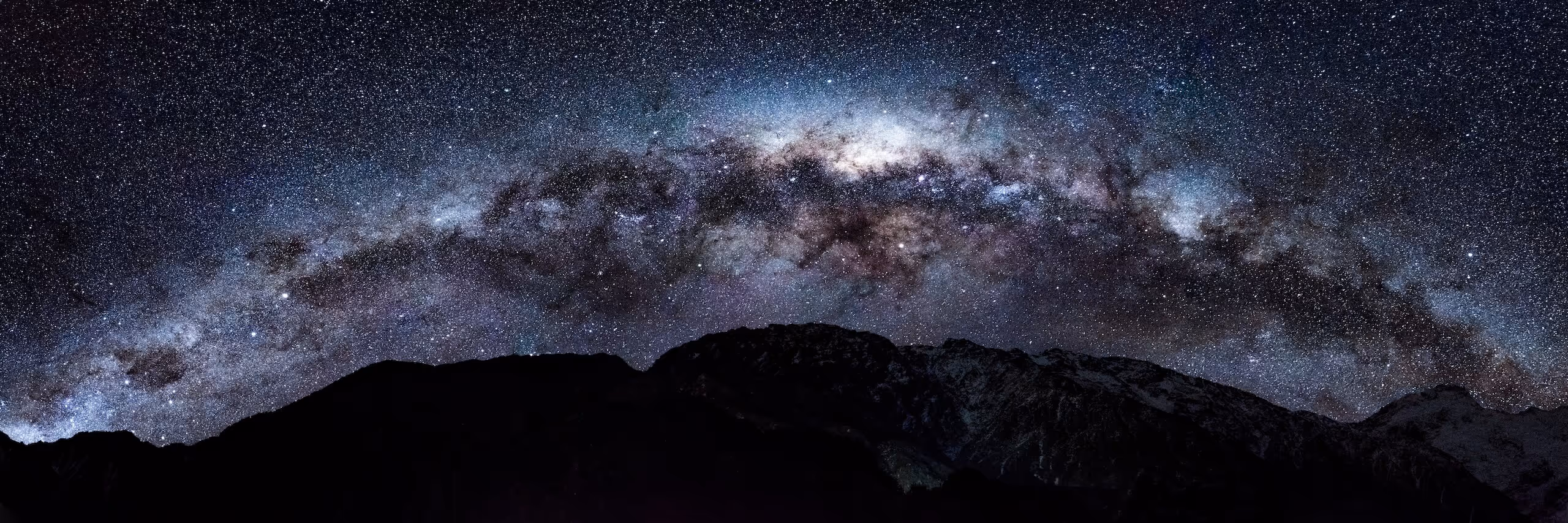 Panoramic view of the Milky Way galaxy arching over dark mountain silhouettes at night.