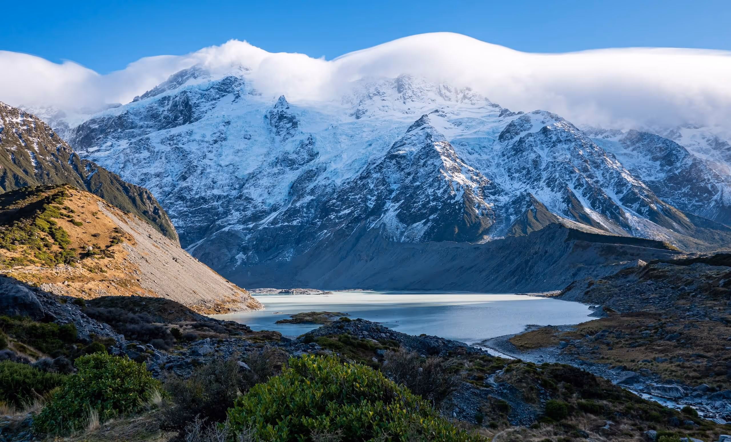 Snow-covered mountain range with clouds at the peaks overlooking a calm alpine lake surrounded by rocky and grassy terrain.
