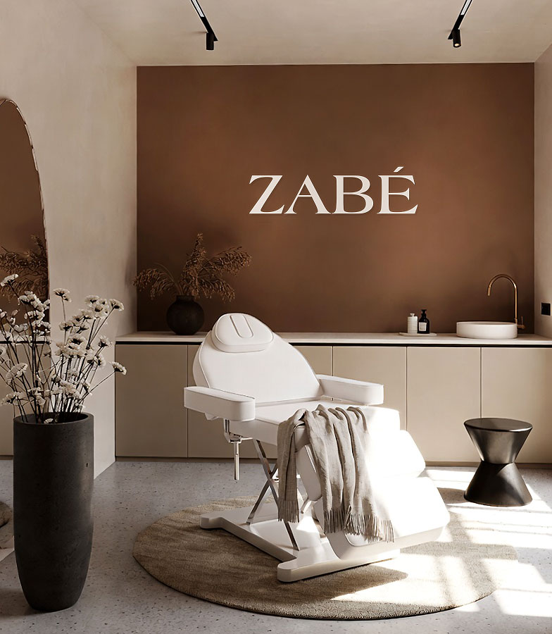 Minimalist spa room with a white reclining chair on a round beige rug, a dark vase with dried flowers, and a brown wall displaying the word ZABÉ.