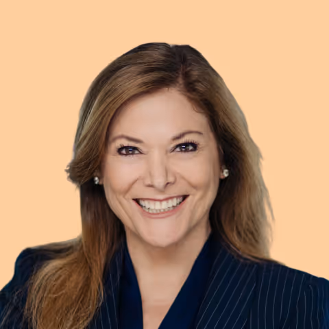 Smiling woman with long light brown hair wearing a dark pinstripe blazer against a peach background.