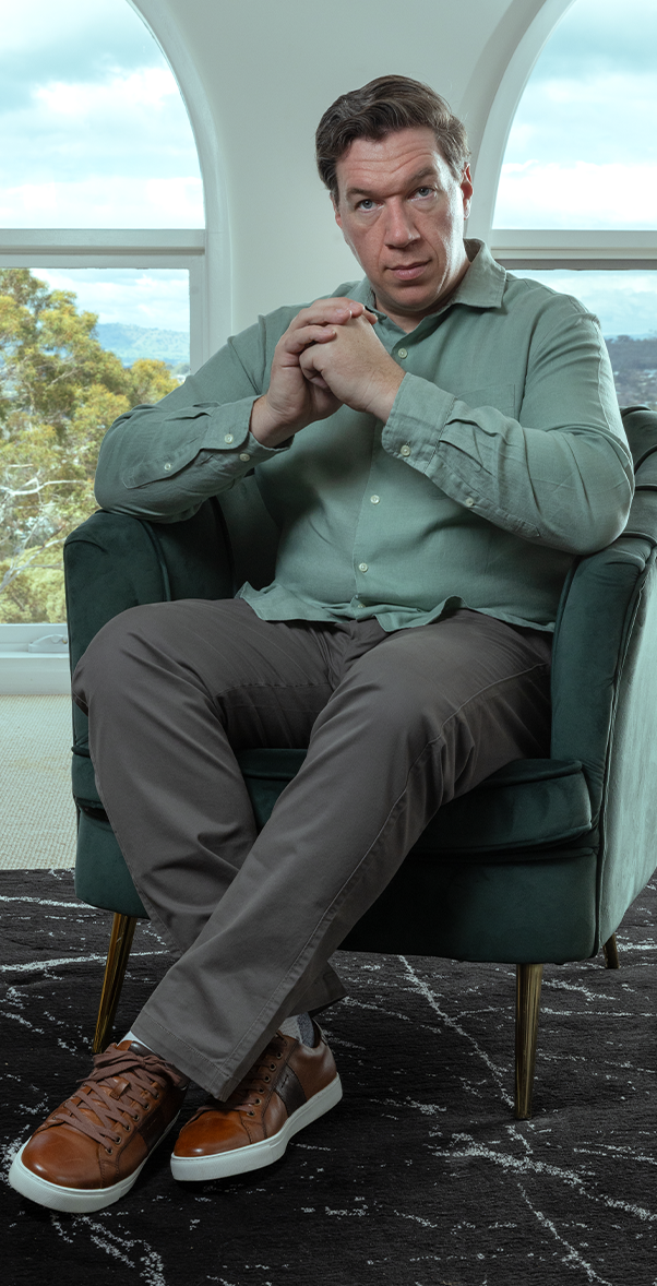 Topher Field standing in a dark jacket against a dramatic studio background, looking directly at the camera with a serious and determined expression.