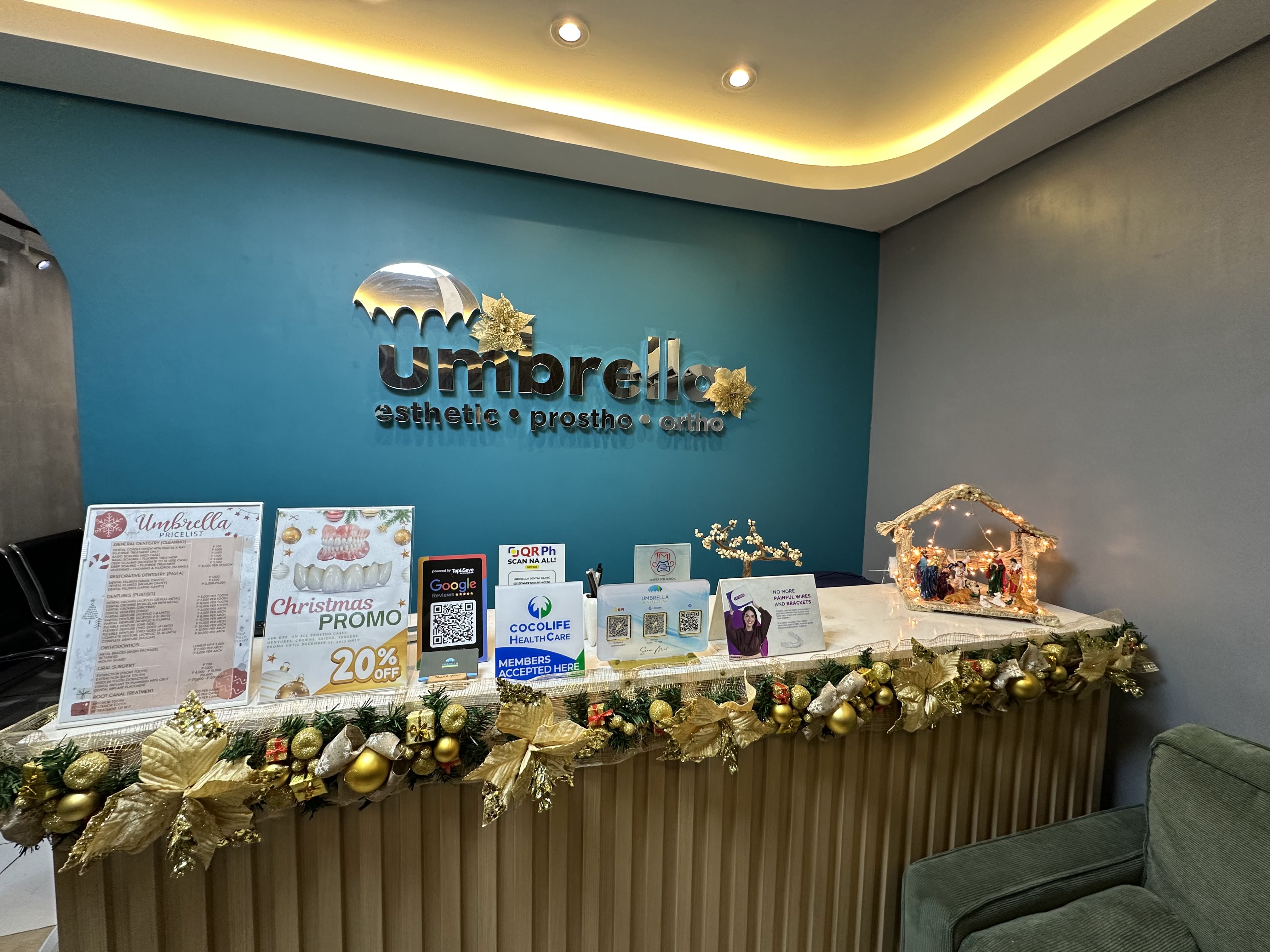 Reception desk of Umbrella Dental Clinic decorated with golden Christmas garland and a lit nativity scene, with Umbrella aesthetic, prostho, and ortho logo on teal wall behind.