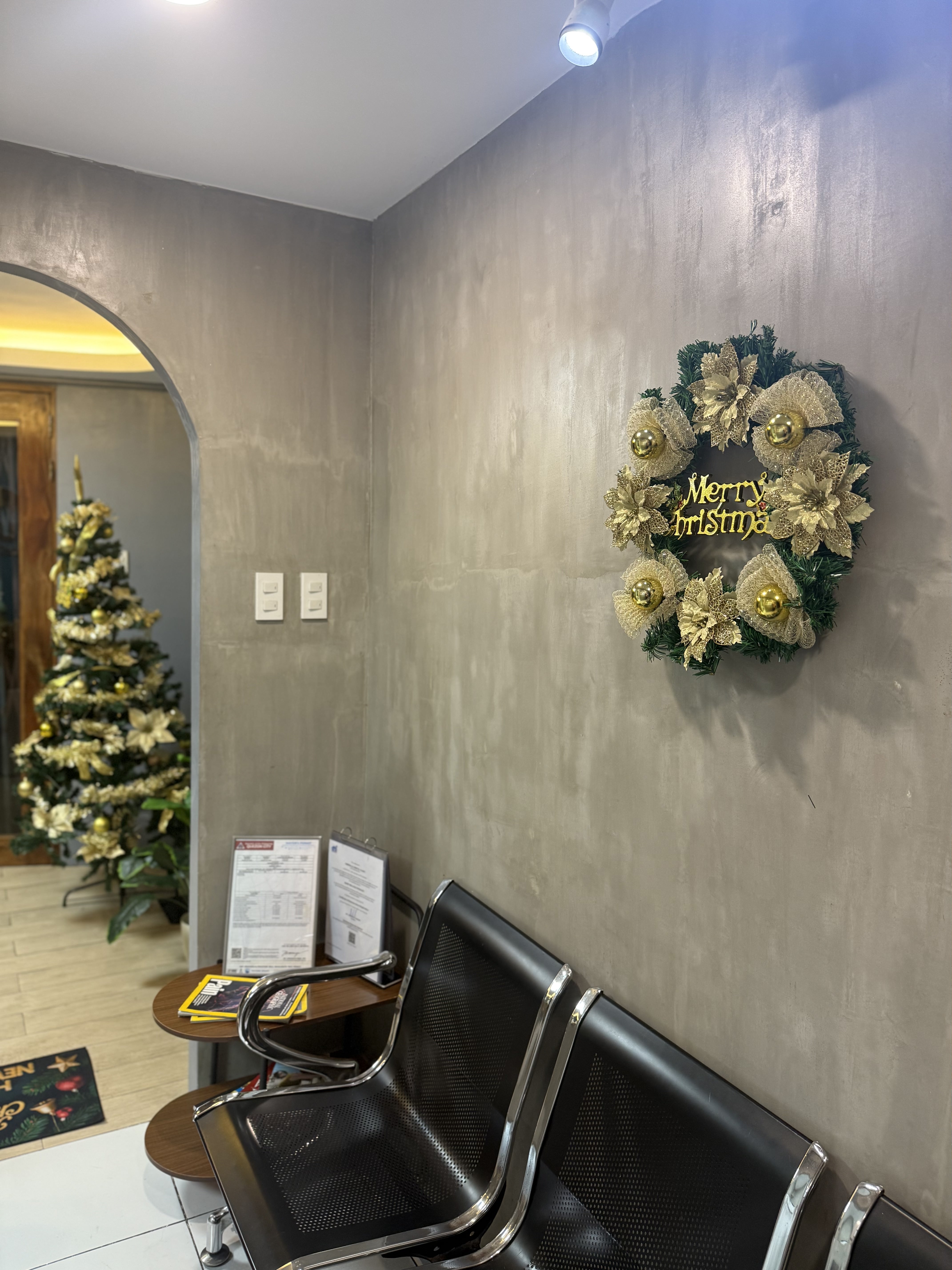 Waiting area of Umbrella Dental Clinic with black metal chairs, wooden side table with documents, a Christmas wreath on a gray wall, and a decorated Christmas tree visible through an archway.