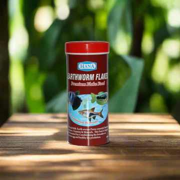Container of HANA Earthworm Flakes premium fish food on a wooden surface with blurred green foliage background.