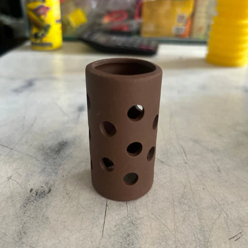 Small cylindrical brown clay object with multiple round holes, placed on a textured white surface.