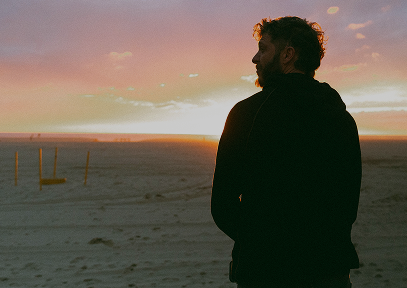 Man in dark clothing looking out over a sandy beach at sunset with a calm sky.