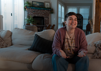 Smiling person with short hair sitting on a beige couch in a cozy living room with a brick fireplace and plants in the background.