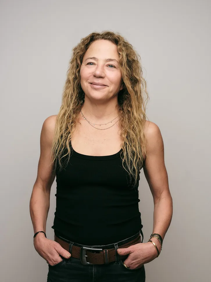 Woman with curly blonde hair wearing a black tank top and jeans, standing with hands in pockets against a neutral background.