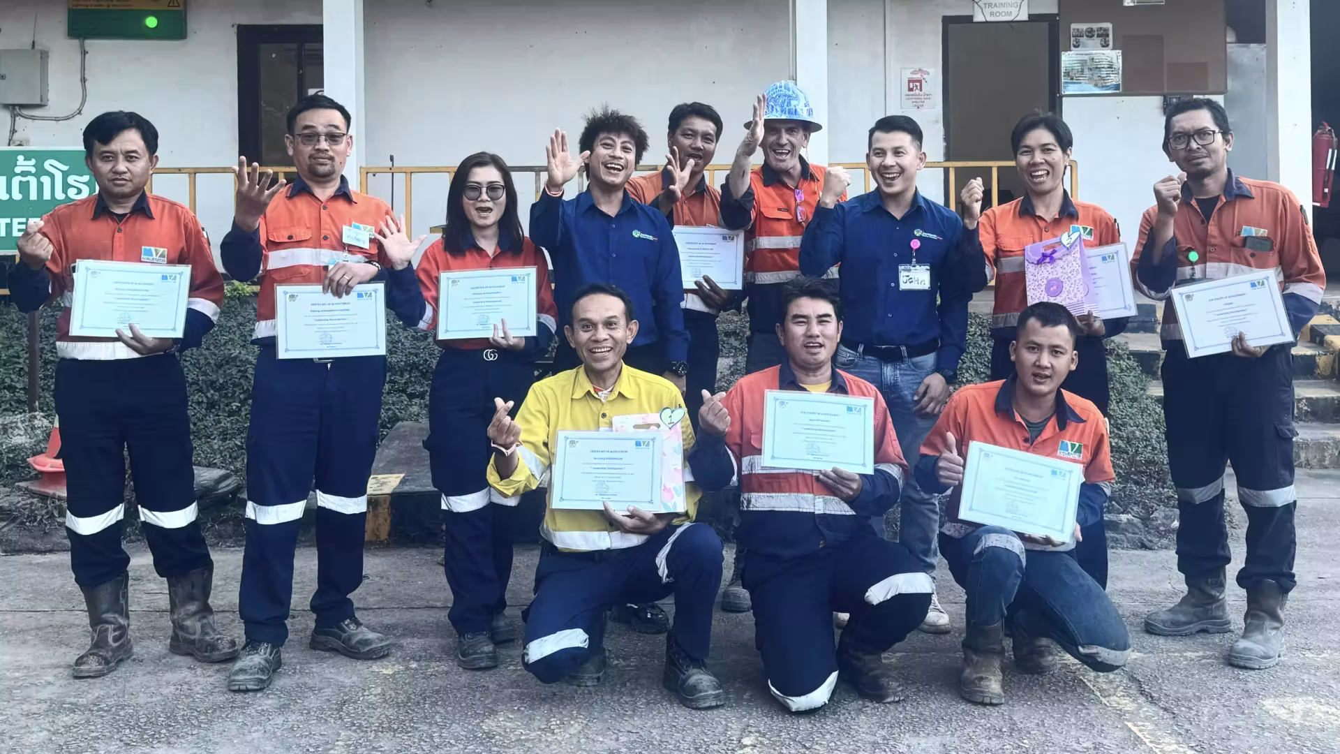 Group of twelve workers in uniforms holding certificates and smiling outdoors in front of a building.