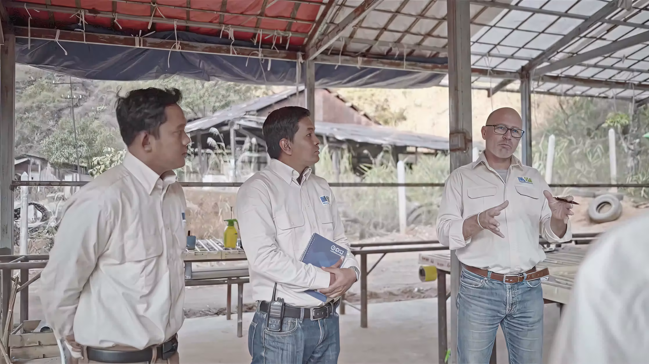 Three men in work shirts and jeans having a discussion in an outdoor covered workspace with rustic buildings and vegetation in the background.