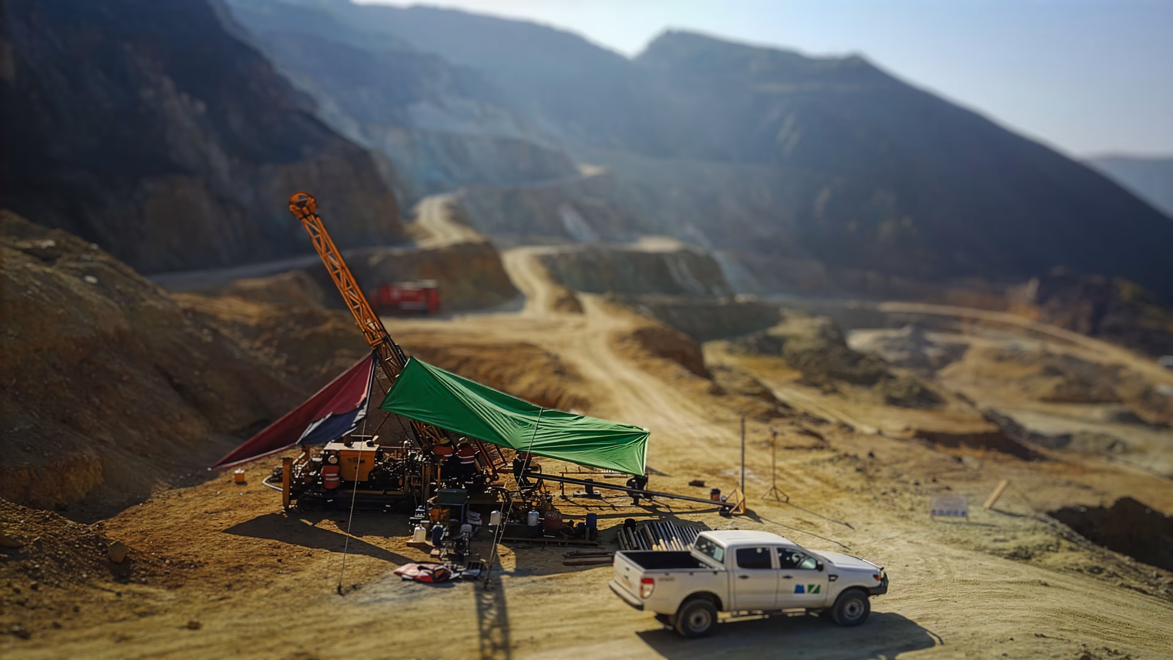 Open-pit mining site with drilling equipment under green and red tarps and a white company pickup truck nearby.