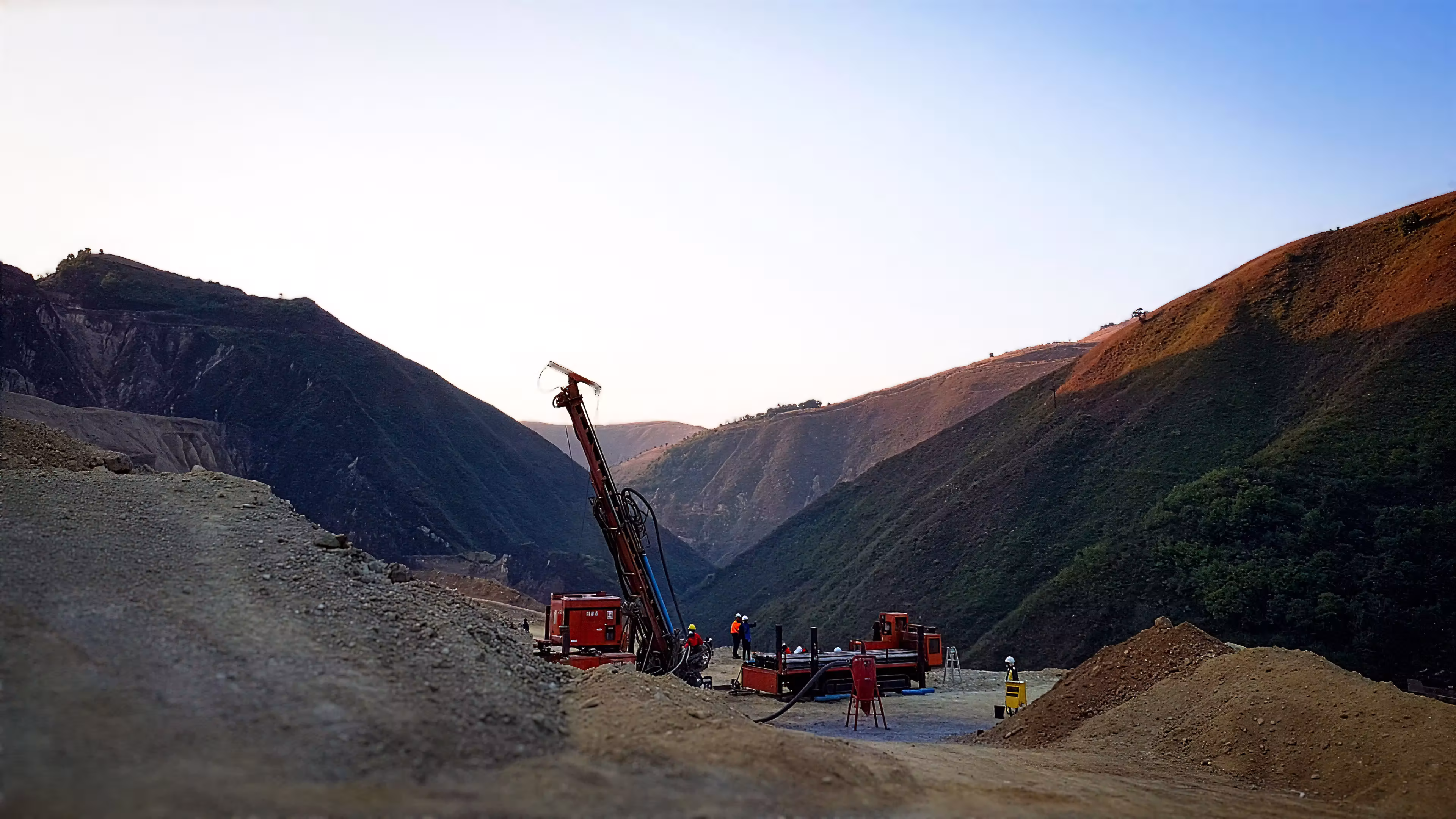 Construction workers operating drilling machinery in a mountainous landscape during sunset.